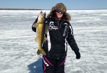 Image of a smiling woman in winter gear standing on a frozen lake holding up a large fish she has caught.